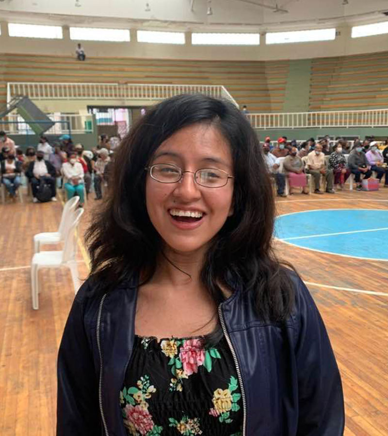 Young woman in Ecuador smiling brightly while wearing new eyeglasses, standing in a gymnasium filled with people.