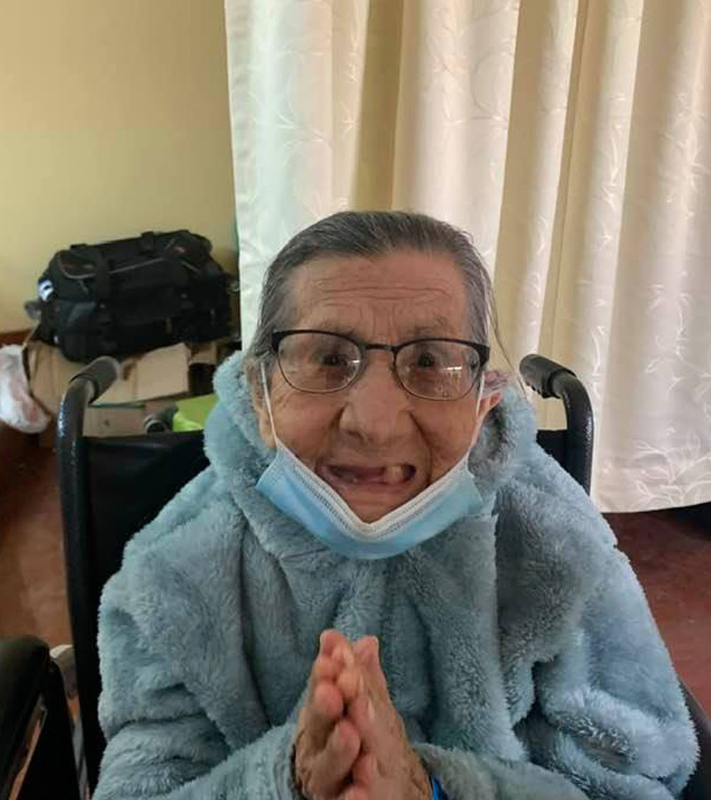 Elderly woman in Peru wearing new eyeglasses, bundled in a blue coat, smiling with hands clasped.